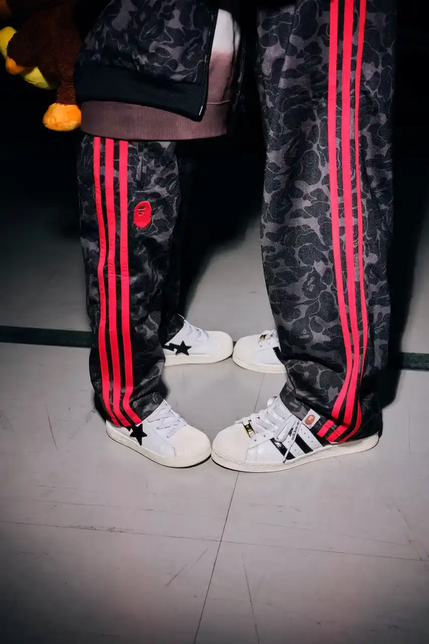 ADIDAS AND BAPE UNITE FOR FRESH COLLECTION AFTER TWO DECADES This article discusses the new collaboration between adidas and A Bathing Ape (BAPE). It details their latest capsule collection centered around the SSTR V BAPE sneaker, which reimagines the classic Superstar silhouette. The article describes the sneaker's design features, available colorways for adults and children, and mentions the accompanying apparel and accessories including tracksuits, hoodies, t-shirts, caps, pouches, and socks. It concludes with information about the April 5th release date through the CONFIRMED app and select retailers. (image 5 of 11)