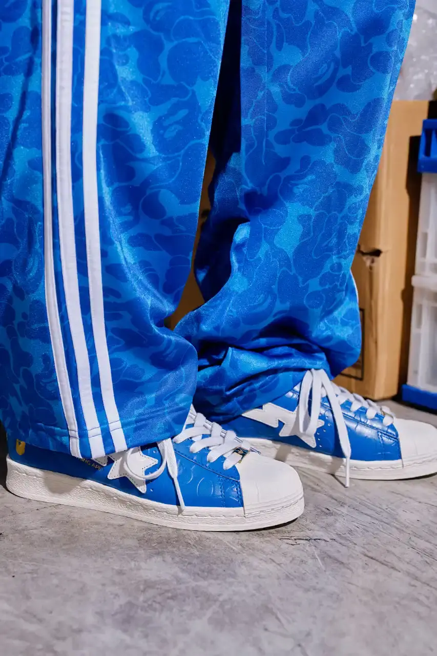 ADIDAS AND BAPE UNITE FOR FRESH COLLECTION AFTER TWO DECADES This article discusses the new collaboration between adidas and A Bathing Ape (BAPE). It details their latest capsule collection centered around the SSTR V BAPE sneaker, which reimagines the classic Superstar silhouette. The article describes the sneaker's design features, available colorways for adults and children, and mentions the accompanying apparel and accessories including tracksuits, hoodies, t-shirts, caps, pouches, and socks. It concludes with information about the April 5th release date through the CONFIRMED app and select retailers. (image 3 of 11)