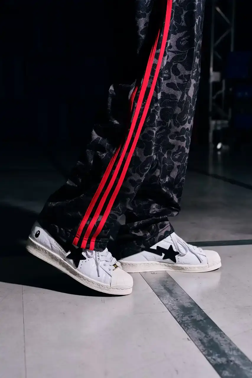 ADIDAS AND BAPE UNITE FOR FRESH COLLECTION AFTER TWO DECADES This article discusses the new collaboration between adidas and A Bathing Ape (BAPE). It details their latest capsule collection centered around the SSTR V BAPE sneaker, which reimagines the classic Superstar silhouette. The article describes the sneaker's design features, available colorways for adults and children, and mentions the accompanying apparel and accessories including tracksuits, hoodies, t-shirts, caps, pouches, and socks. It concludes with information about the April 5th release date through the CONFIRMED app and select retailers. (image 4 of 11)