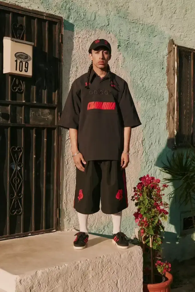 ADIDAS ORIGINALS TEAMS WITH WILLY CHAVARRIA ON CHICANO-INSPIRED COLLECTION image of the Spring/Summer 2025 collaboration between adidas Originals and designer Willy Chavarria. The collection celebrates Chicano culture through fashion, featuring oversized silhouettes and Mexican cultural symbols. Key pieces include tracksuits with Three Stripes, redesigned Trefoil logos with rose embroidery, co-branded hoodies with red and green panels, and the revamped 1978 Jabbar basketball shoe. The collection launches in two drops: May 8 and July 10, available through the CONFIRMED app and select retailers. The campaign was photographed by Carlos Jaramillo in Los Angeles with Chavarria's friends and family. (image 7 of 10)