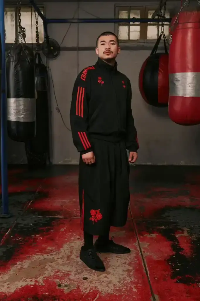 ADIDAS ORIGINALS TEAMS WITH WILLY CHAVARRIA ON CHICANO-INSPIRED COLLECTION image of the Spring/Summer 2025 collaboration between adidas Originals and designer Willy Chavarria. The collection celebrates Chicano culture through fashion, featuring oversized silhouettes and Mexican cultural symbols. Key pieces include tracksuits with Three Stripes, redesigned Trefoil logos with rose embroidery, co-branded hoodies with red and green panels, and the revamped 1978 Jabbar basketball shoe. The collection launches in two drops: May 8 and July 10, available through the CONFIRMED app and select retailers. The campaign was photographed by Carlos Jaramillo in Los Angeles with Chavarria's friends and family. (image 5 of 10)