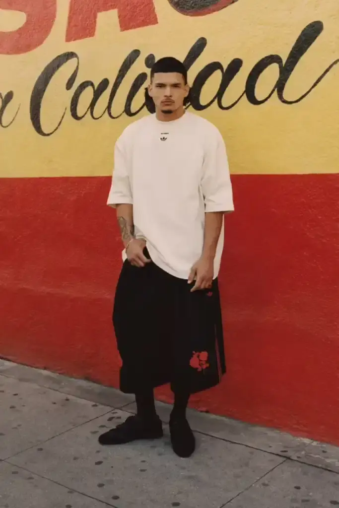 ADIDAS ORIGINALS TEAMS WITH WILLY CHAVARRIA ON CHICANO-INSPIRED COLLECTION image of the Spring/Summer 2025 collaboration between adidas Originals and designer Willy Chavarria. The collection celebrates Chicano culture through fashion, featuring oversized silhouettes and Mexican cultural symbols. Key pieces include tracksuits with Three Stripes, redesigned Trefoil logos with rose embroidery, co-branded hoodies with red and green panels, and the revamped 1978 Jabbar basketball shoe. The collection launches in two drops: May 8 and July 10, available through the CONFIRMED app and select retailers. The campaign was photographed by Carlos Jaramillo in Los Angeles with Chavarria's friends and family. (image 2 of 10)