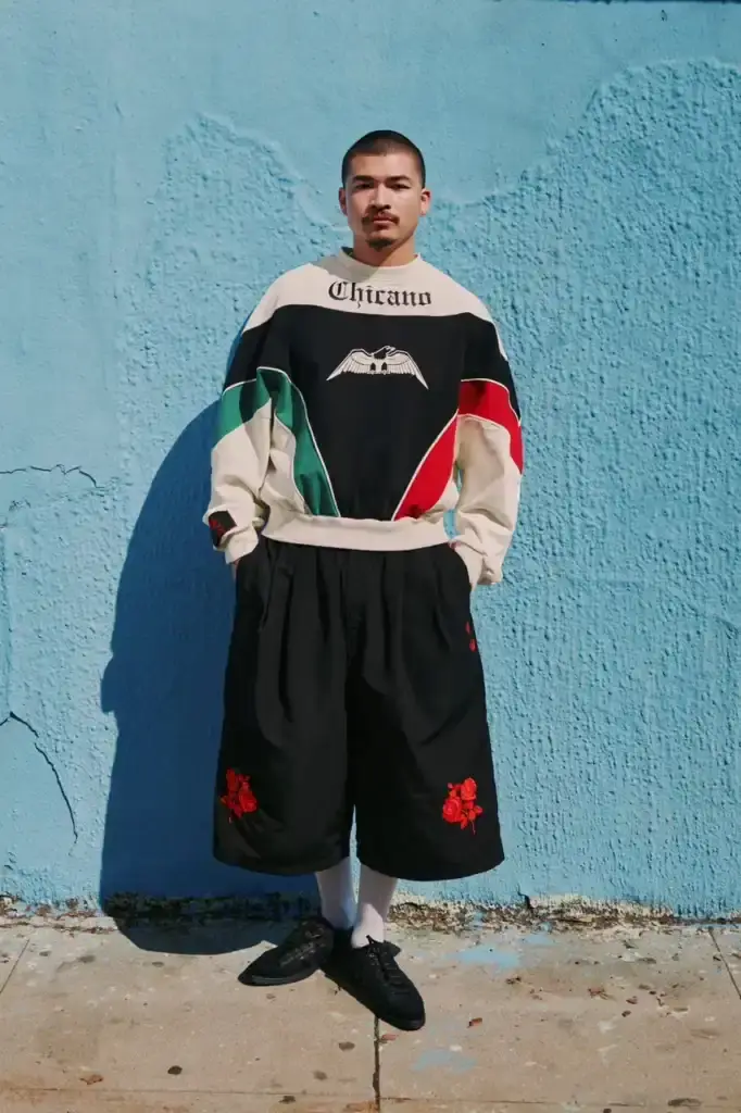 ADIDAS ORIGINALS TEAMS WITH WILLY CHAVARRIA ON CHICANO-INSPIRED COLLECTION image of the Spring/Summer 2025 collaboration between adidas Originals and designer Willy Chavarria. The collection celebrates Chicano culture through fashion, featuring oversized silhouettes and Mexican cultural symbols. Key pieces include tracksuits with Three Stripes, redesigned Trefoil logos with rose embroidery, co-branded hoodies with red and green panels, and the revamped 1978 Jabbar basketball shoe. The collection launches in two drops: May 8 and July 10, available through the CONFIRMED app and select retailers. The campaign was photographed by Carlos Jaramillo in Los Angeles with Chavarria's friends and family. (image 3 of 10)