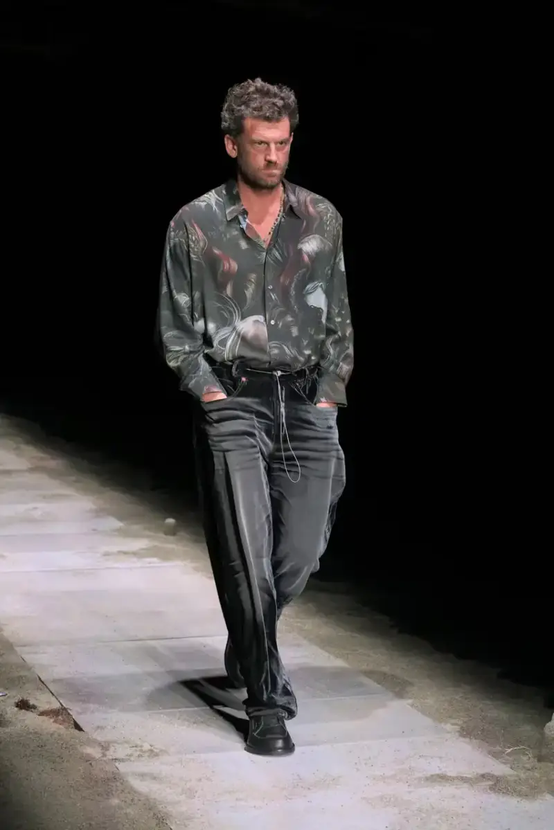 Magliano Models walk on sand-covered runway inside dark gymnasium, wearing layered winter clothing with misaligned buttons and multiple belts from Magliano's FW25 collection. (image 7 of 35)