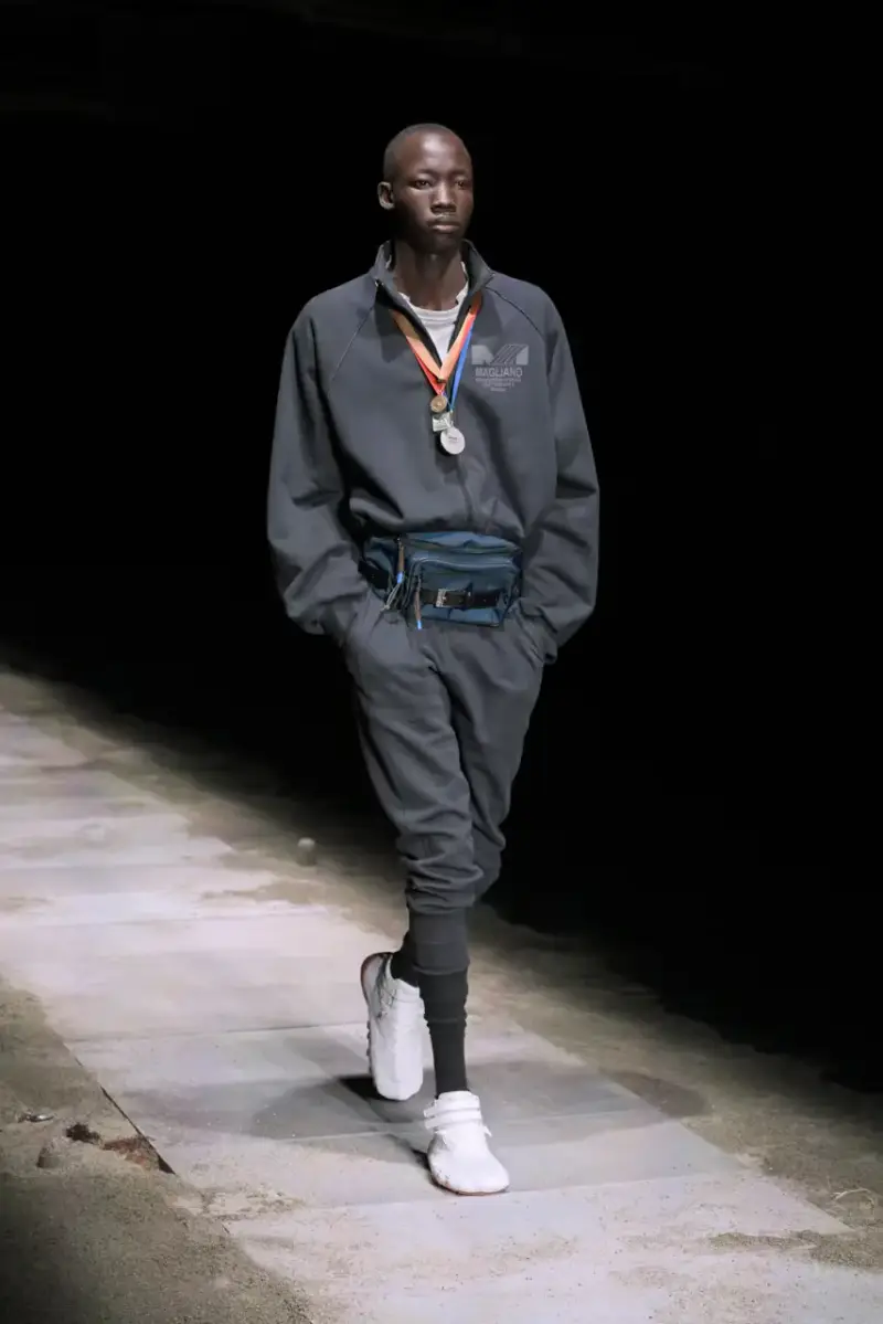 Magliano Models walk on sand-covered runway inside dark gymnasium, wearing layered winter clothing with misaligned buttons and multiple belts from Magliano's FW25 collection. (image 9 of 35)