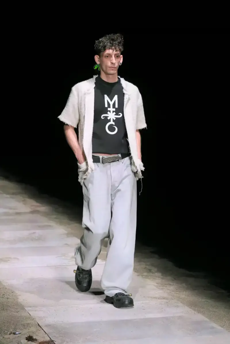 Magliano Models walk on sand-covered runway inside dark gymnasium, wearing layered winter clothing with misaligned buttons and multiple belts from Magliano's FW25 collection. (image 5 of 35)