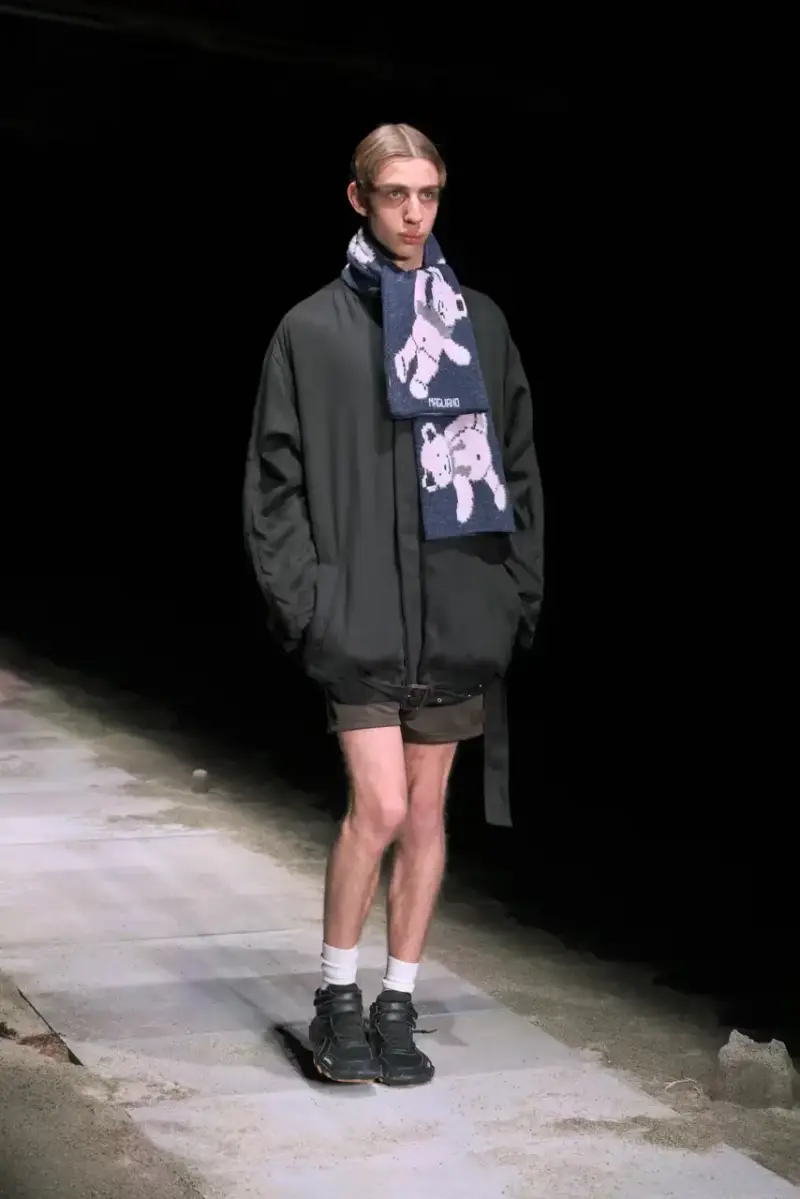 Magliano Models walk on sand-covered runway inside dark gymnasium, wearing layered winter clothing with misaligned buttons and multiple belts from Magliano's FW25 collection. (image 8 of 35)