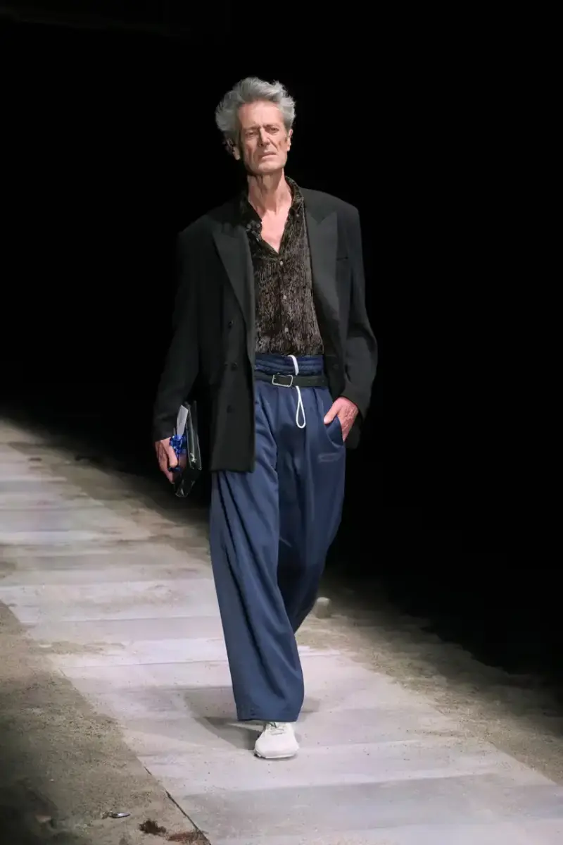 Magliano Models walk on sand-covered runway inside dark gymnasium, wearing layered winter clothing with misaligned buttons and multiple belts from Magliano's FW25 collection. (image 6 of 35)