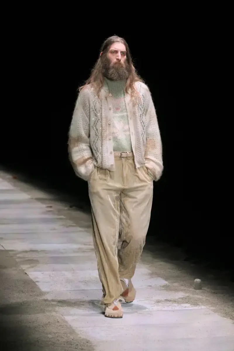 Magliano Models walk on sand-covered runway inside dark gymnasium, wearing layered winter clothing with misaligned buttons and multiple belts from Magliano's FW25 collection. (image 19 of 35)