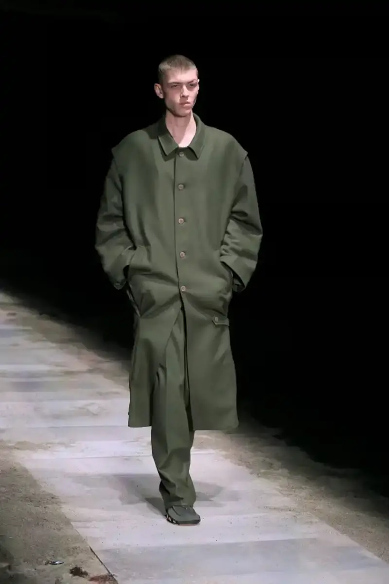 Magliano Models walk on sand-covered runway inside dark gymnasium, wearing layered winter clothing with misaligned buttons and multiple belts from Magliano's FW25 collection. (image 34 of 35)