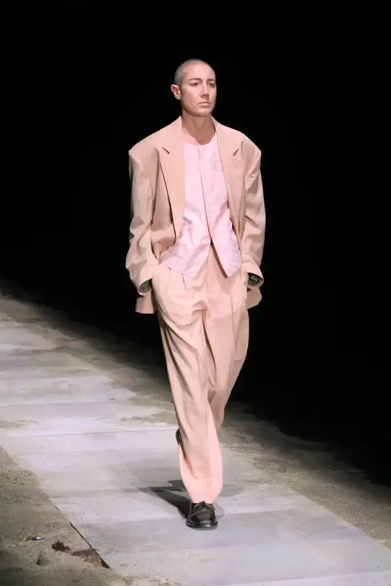 Magliano Models walk on sand-covered runway inside dark gymnasium, wearing layered winter clothing with misaligned buttons and multiple belts from Magliano's FW25 collection. (image 2 of 35)