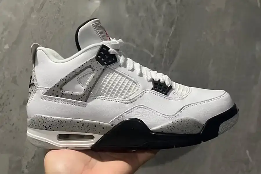 Air Jordan 4 sneaker in White Cement colorway, showing white leather upper with speckled grey overlays, black and red accents, visible Air unit in heel, and Nike Air branding on heel tab. The sneaker is photographed from a side angle on a neutral background.