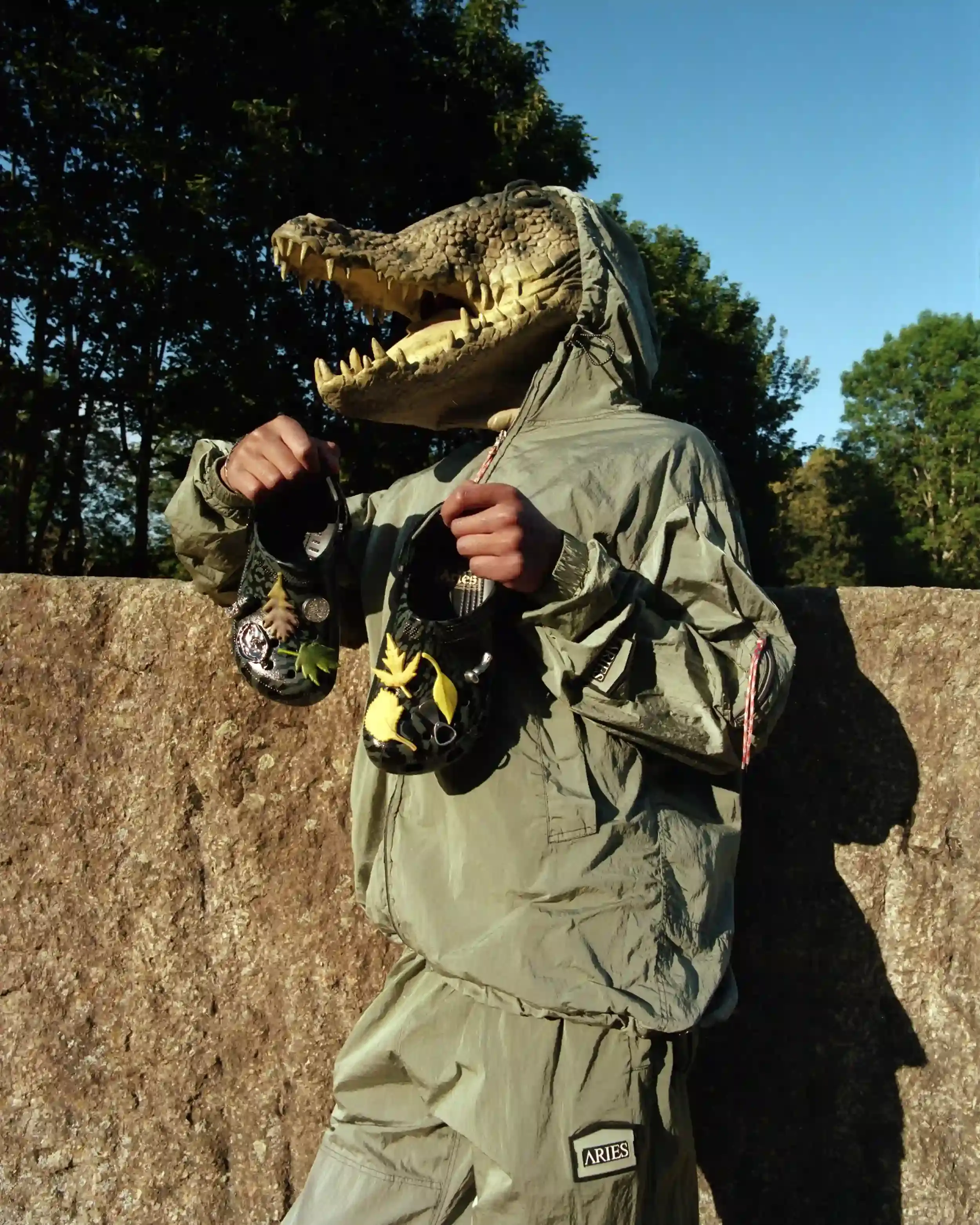 ARIES RETURNS WITH SECOND CROCS COLLECTION image about the second collaboration between London streetwear brand Aries and footwear company Crocs. The collection features two camouflage-patterned Classic Clogs in High Shine Black and Dark Algae Green colorways, accompanied by custom Jibbitz charms inspired by urban artifacts. The products launch July 23, 2025, priced at £75.00, available through Aries' website, flagship store, Crocs app, and select retailers worldwide.