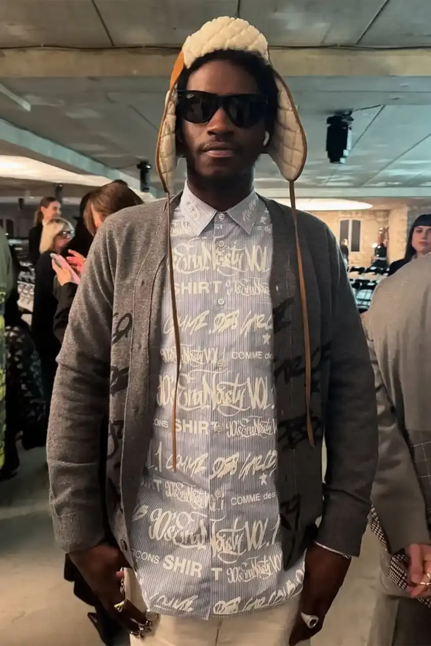 A$AP Nast at Paris Fashion Week wearing a blue striped oxford shirt featuring AWGE, Opium, and COMME des GARÇONS SHIRT logos, layered over a grey cardigan with graffiti-style artwork.