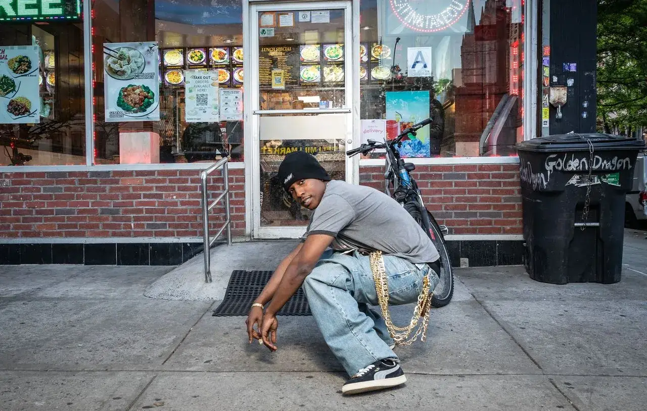 ASAP ROCKY RECREATES PUMA SUEDE 94 SNEAKER person squats on a New York sidewalk wearing baggy jeans, a grey shirt, a black beanie, and black and white Puma Suede sneakers. A bicycle and a shop entrance stand behind the person.