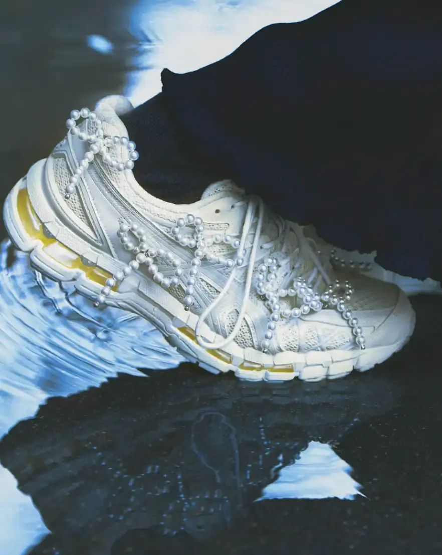 ASICS AND COMPLETEDWORKS RELEASE GEL-KAYANO 20 COLLABORATION A photograph shows a pair of ASICS GEL-KAYANO 20 sneakers modified with sculptural jewelry elements like pearls and floral motifs.