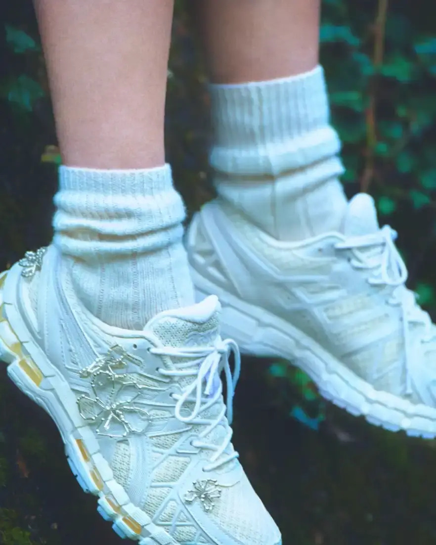 ASICS AND COMPLETEDWORKS RELEASE GEL-KAYANO 20 COLLABORATION A photograph shows a pair of ASICS GEL-KAYANO 20 sneakers modified with sculptural jewelry elements like pearls and floral motifs. (image 5 of 6)