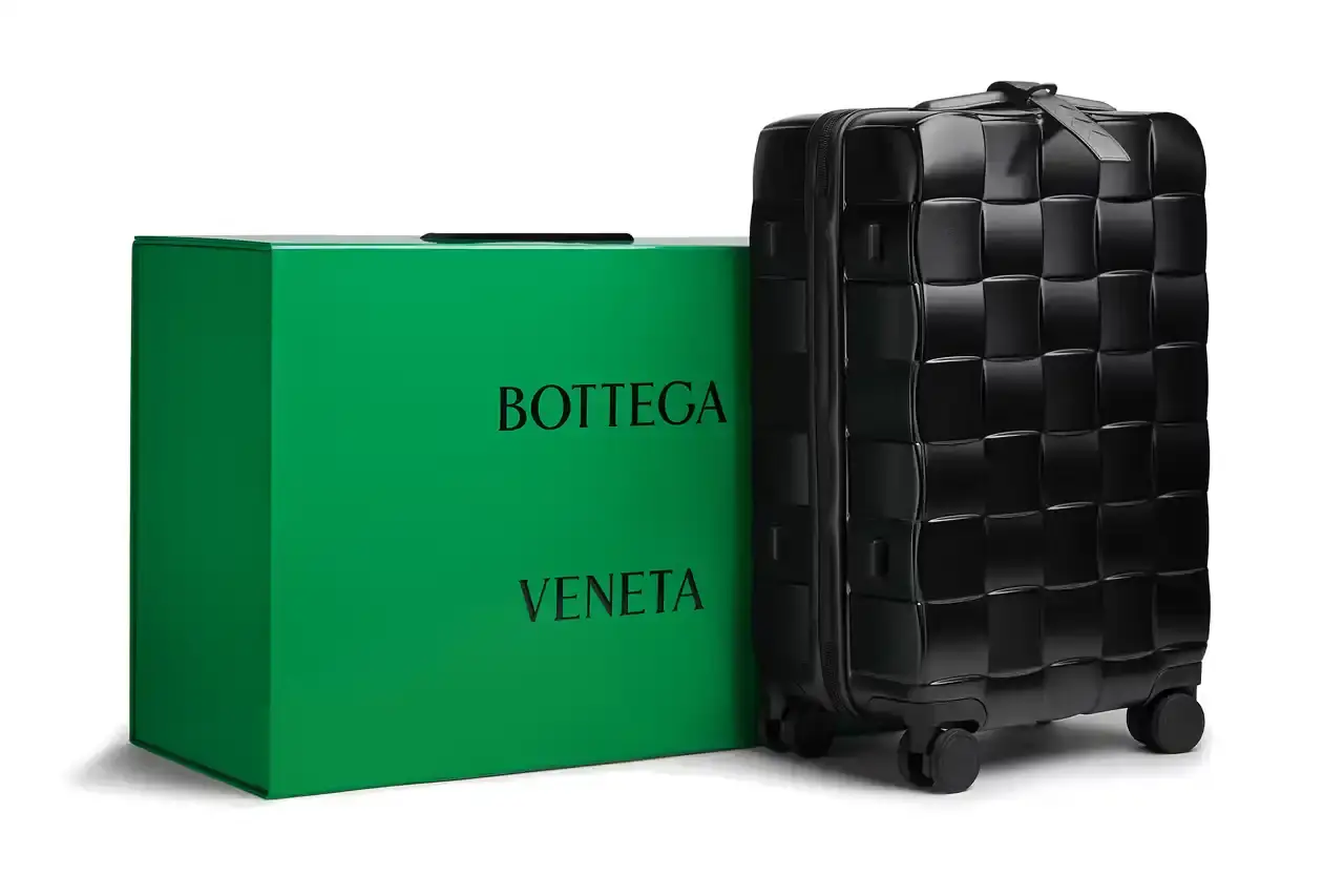 Bottega Veneta Bottega Veneta Odyssey carry-on suitcase with textured polycarbonate shell featuring 3D Intreccio pattern, silver hardware, and brown calfskin leather handles. The wheeled luggage is shown in a three-quarter view displaying its structured design and branded details.