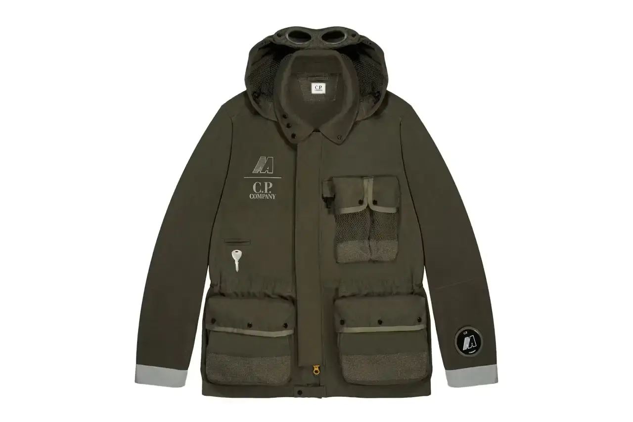 Automobili Amos The C.P. Company x Automobili Amos Mille Jacket shown in signature green color, featuring the distinctive goggle hood design, reflective details, and technical mesh construction. The jacket is photographed against a neutral background, displaying its motorsport-inspired details including the watch viewer window and specialized key pocket.