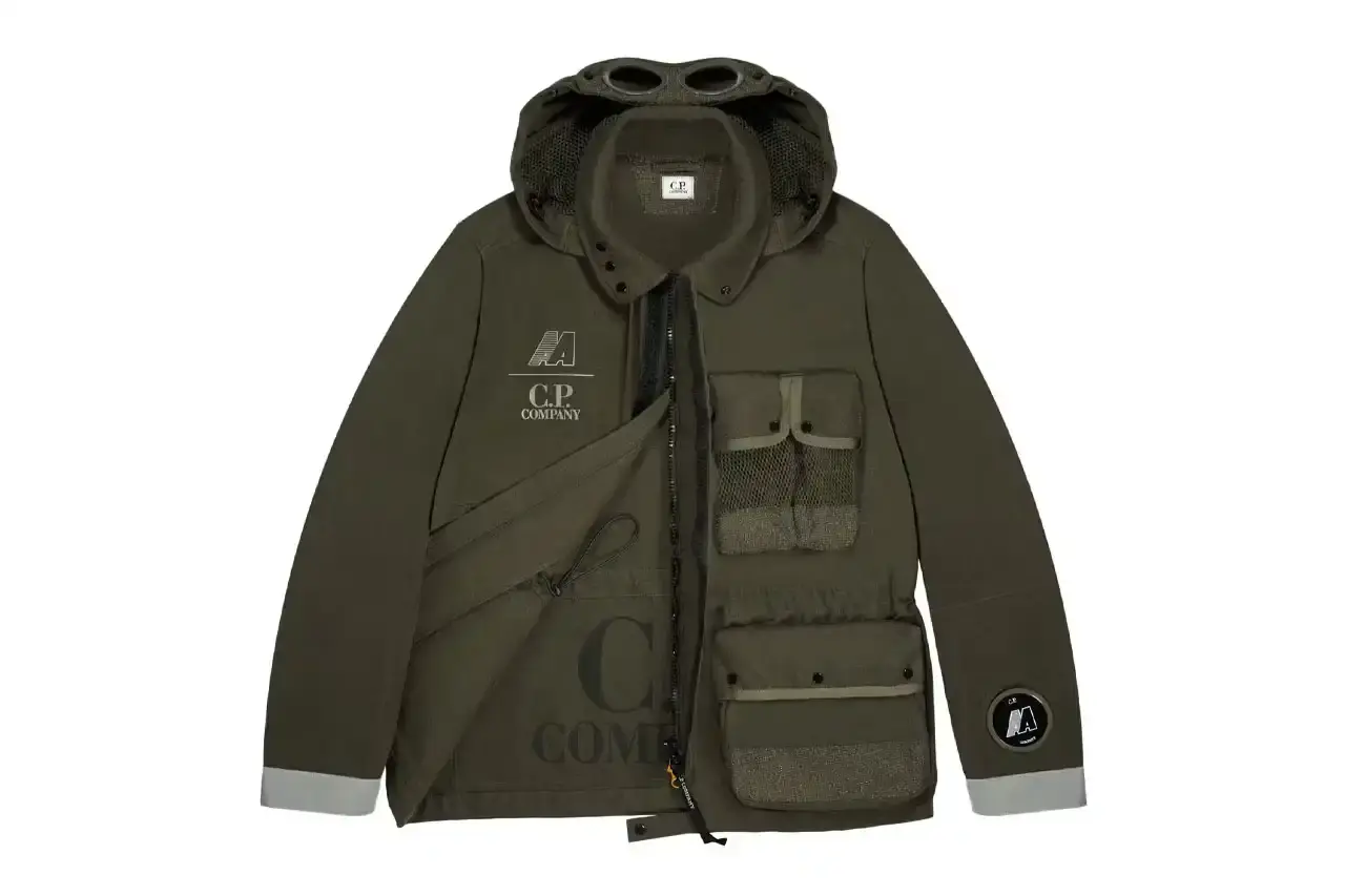 Automobili Amos The C.P. Company x Automobili Amos Mille Jacket shown in signature green color, featuring the distinctive goggle hood design, reflective details, and technical mesh construction. The jacket is photographed against a neutral background, displaying its motorsport-inspired details including the watch viewer window and specialized key pocket. (image 3 of 10)