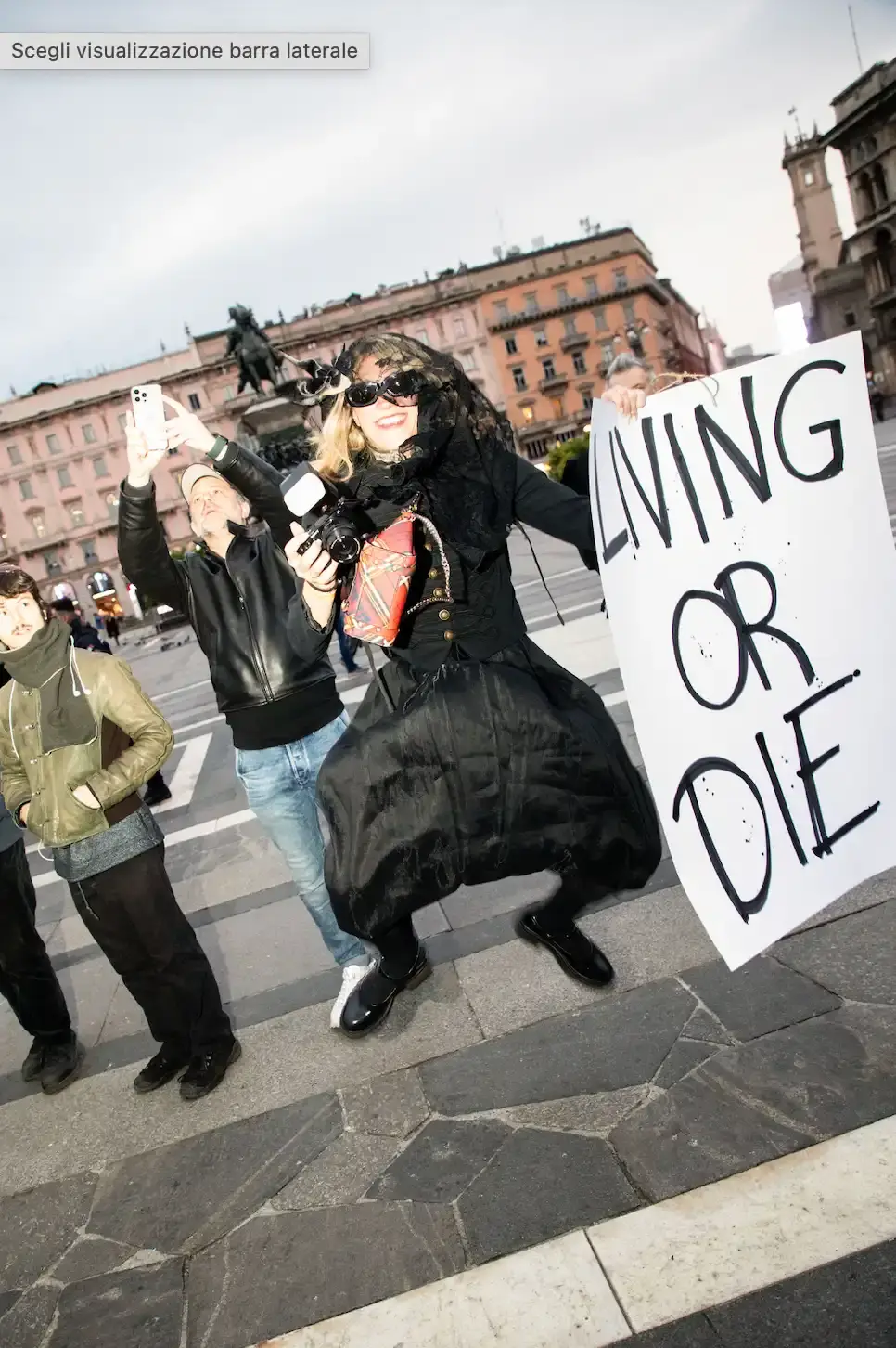 CATTELAN AND BALLARIO TRANSFORM GRIEF INTO DAWN PERFORMANCE IN MILAN'S DUOMO SQUARE Photo of an art performance called "The Cheerful End" organized by artist Maurizio Cattelan and critic Nicolas Ballario in Milan. Held at dawn in Piazza Duomo, participants displayed humorous and philosophical signs about death. The event closed Milan Art Week and was supported by the Museo del Novecento. The performance used irony to encourage public reflection on mortality, with refreshments moved indoors at authorities' request. The article describes how the two creators acted as "accomplices in absurdity" to transform a somber subject into a thought-provoking shared experience. (image 8 of 10)
