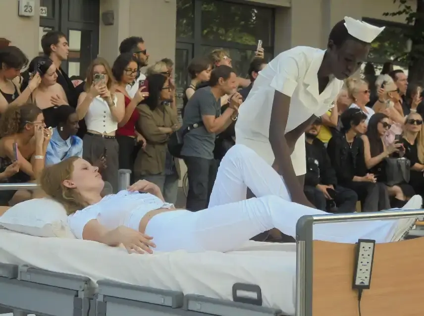 image of Clara Colette Miramon's SS26 "CARE" collection presented at Berlin Fashion Week. The innovative show featured models emerging from an ambulance, hospital bed installations, and Pilates reformer performances to highlight the physical and psychological demands placed on women in caregiving roles. The presentation combined fashion with powerful social commentary about making women's invisible labor visible through strategic cut-out designs, medical equipment accessories, and an immersive installation format that transformed the runway into a statement about women's multifaceted roles in society.