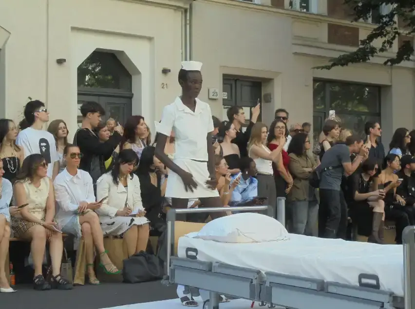 image of Clara Colette Miramon's SS26 "CARE" collection presented at Berlin Fashion Week. The innovative show featured models emerging from an ambulance, hospital bed installations, and Pilates reformer performances to highlight the physical and psychological demands placed on women in caregiving roles. The presentation combined fashion with powerful social commentary about making women's invisible labor visible through strategic cut-out designs, medical equipment accessories, and an immersive installation format that transformed the runway into a statement about women's multifaceted roles in society. (image 3 of 4)