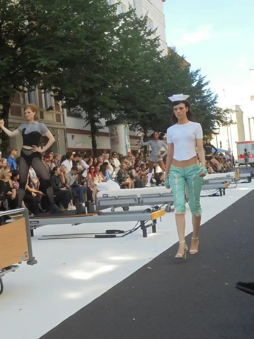 image of Clara Colette Miramon's SS26 "CARE" collection presented at Berlin Fashion Week. The innovative show featured models emerging from an ambulance, hospital bed installations, and Pilates reformer performances to highlight the physical and psychological demands placed on women in caregiving roles. The presentation combined fashion with powerful social commentary about making women's invisible labor visible through strategic cut-out designs, medical equipment accessories, and an immersive installation format that transformed the runway into a statement about women's multifaceted roles in society. (image 4 of 4)