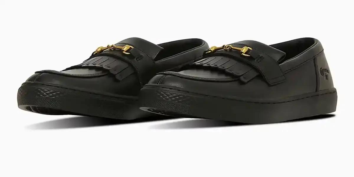 Converse Converse All-Star Coupe GB Loafer shown from side angle, featuring minimalist leather construction, tasseled midfoot strap with embossed star details, and classic Converse sole. Available in black and off-white colorways.