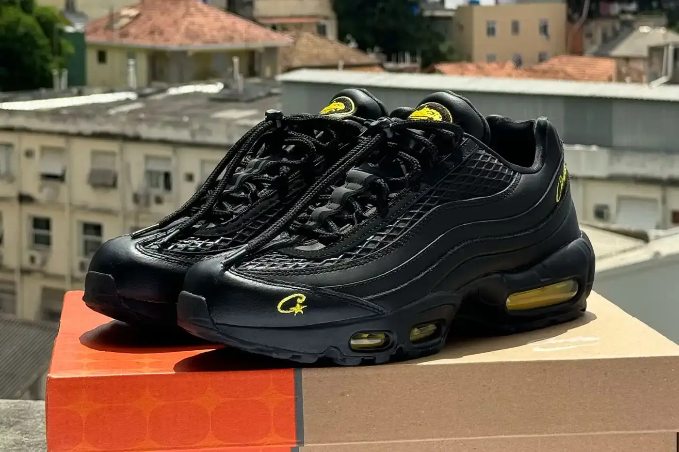 Clint419 Corteiz x Nike Air Max 95 'Honey Black' sneaker featuring an all-black premium leather upper with contrasting Tour Yellow accents on the branding and Air cushioning units. The sleek design showcases Corteiz branding on the lateral toe, heel, tongue and outsole, with London coordinates visible on the heel. The shoe maintains the Air Max 95's signature layered construction with mesh detailing on the upper.