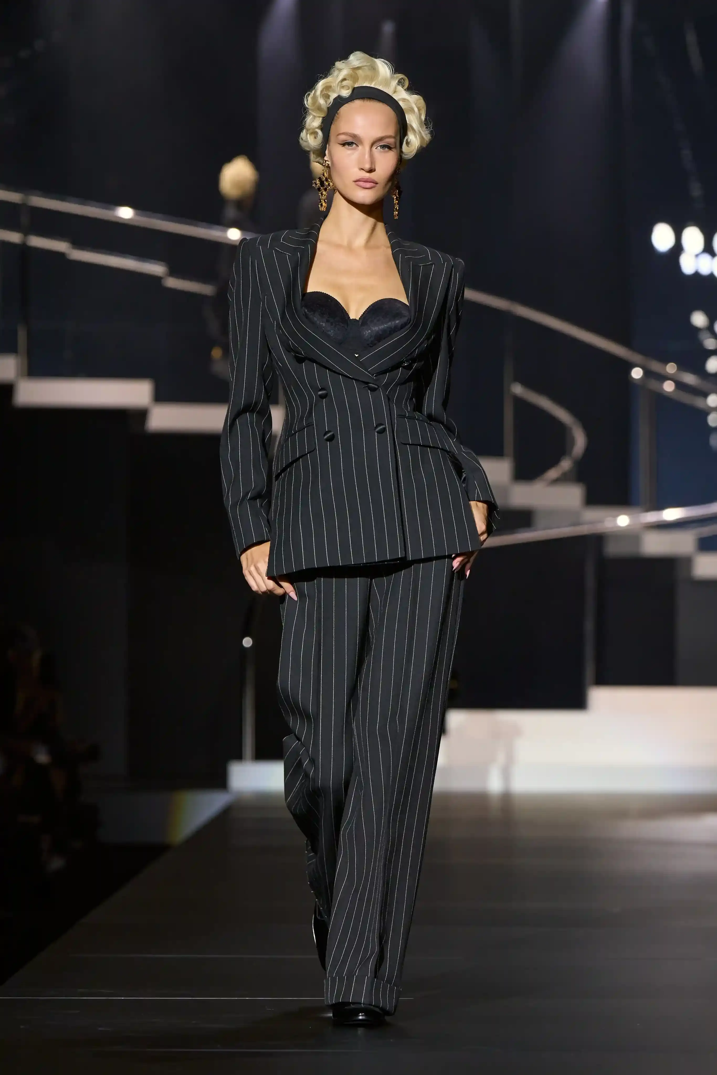 Dolce & Gabbana's Spring/Summer 2025 runway show at Milan Fashion Week showcases a collection inspired by Madonna's iconic style. The presentation features body-contouring designs, including anatomically cut jackets and corsetry details, in materials such as duchesse satin, crepe, tulle, chiffon, lace, and faux fur. The color palette focuses on black, nude, red, and white, with notable pieces including conical bras and oversized cross earrings - signatures of Madonna's most memorable looks. Attended by celebrities like Naomi Campbell and Ashley Graham, the show celebrates Madonna's enduring influence on fashion and her ability to challenge societal norms while remaining true to herself. This collection reinforces Dolce & Gabbana's commitment to empowering women's fashion and highlights the lasting impact of cultural icons on design (image 8 of 9)