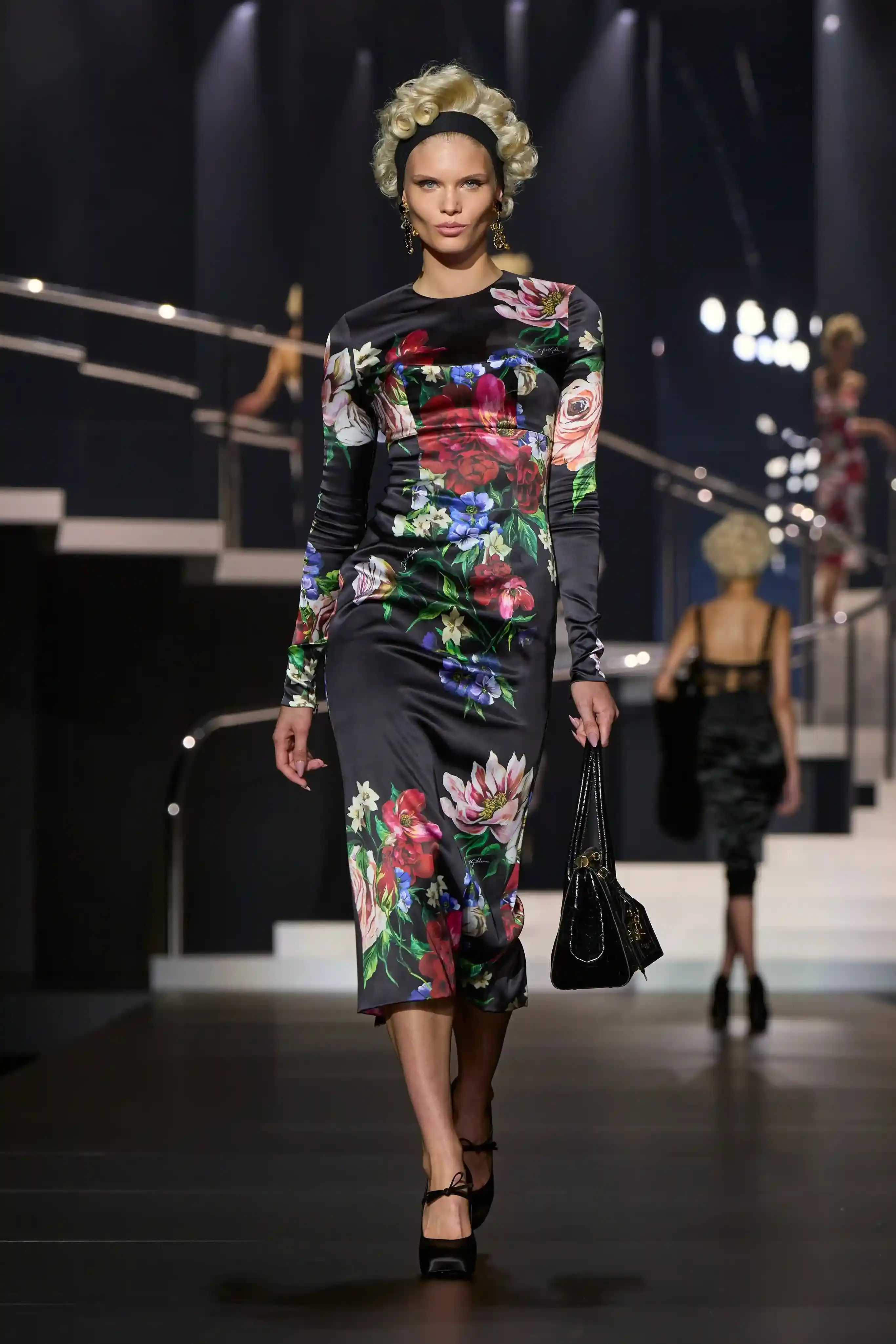 Dolce & Gabbana's Spring/Summer 2025 runway show at Milan Fashion Week showcases a collection inspired by Madonna's iconic style. The presentation features body-contouring designs, including anatomically cut jackets and corsetry details, in materials such as duchesse satin, crepe, tulle, chiffon, lace, and faux fur. The color palette focuses on black, nude, red, and white, with notable pieces including conical bras and oversized cross earrings - signatures of Madonna's most memorable looks. Attended by celebrities like Naomi Campbell and Ashley Graham, the show celebrates Madonna's enduring influence on fashion and her ability to challenge societal norms while remaining true to herself. This collection reinforces Dolce & Gabbana's commitment to empowering women's fashion and highlights the lasting impact of cultural icons on design (image 6 of 9)