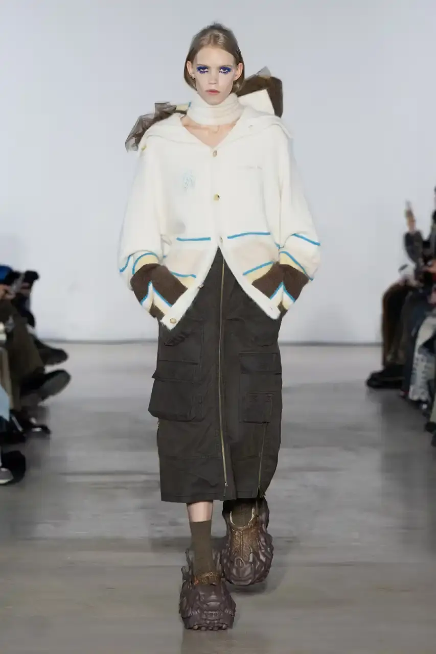 Feng Chen Wang Runway image of a model wearing Feng Chen Wang's Fall/Winter 2025 collection, showcasing oversized ceramic-textured UGG slippers and layered outerwear inspired by mythical Chinese creatures from the Shan Hai Jing text (image 6 of 11)