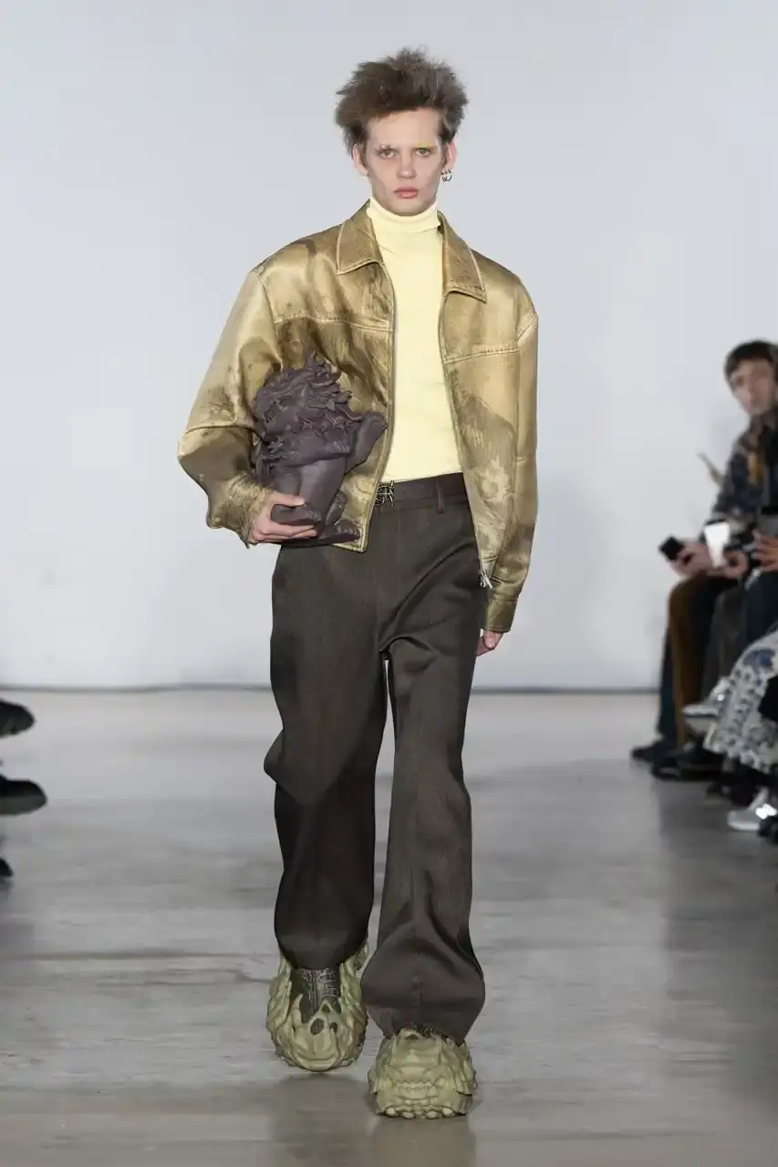 Feng Chen Wang Runway image of a model wearing Feng Chen Wang's Fall/Winter 2025 collection, showcasing oversized ceramic-textured UGG slippers and layered outerwear inspired by mythical Chinese creatures from the Shan Hai Jing text (image 11 of 11)