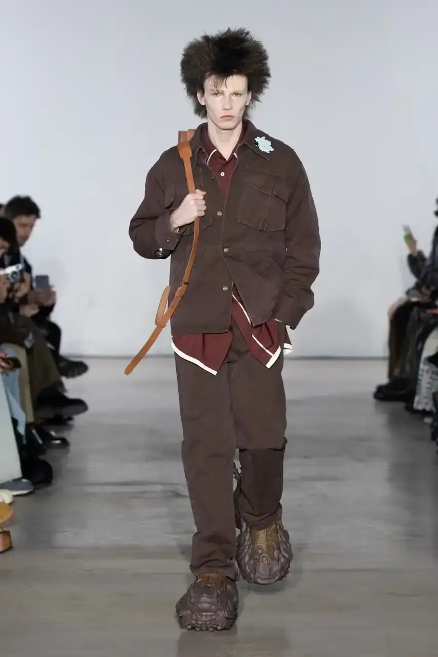Feng Chen Wang Runway image of a model wearing Feng Chen Wang's Fall/Winter 2025 collection, showcasing oversized ceramic-textured UGG slippers and layered outerwear inspired by mythical Chinese creatures from the Shan Hai Jing text (image 7 of 11)
