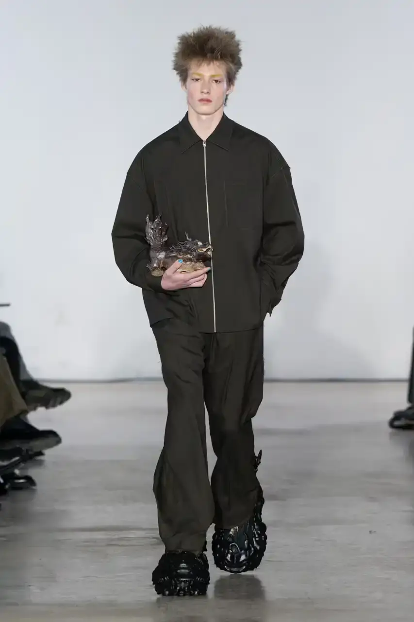 Feng Chen Wang Runway image of a model wearing Feng Chen Wang's Fall/Winter 2025 collection, showcasing oversized ceramic-textured UGG slippers and layered outerwear inspired by mythical Chinese creatures from the Shan Hai Jing text (image 3 of 11)