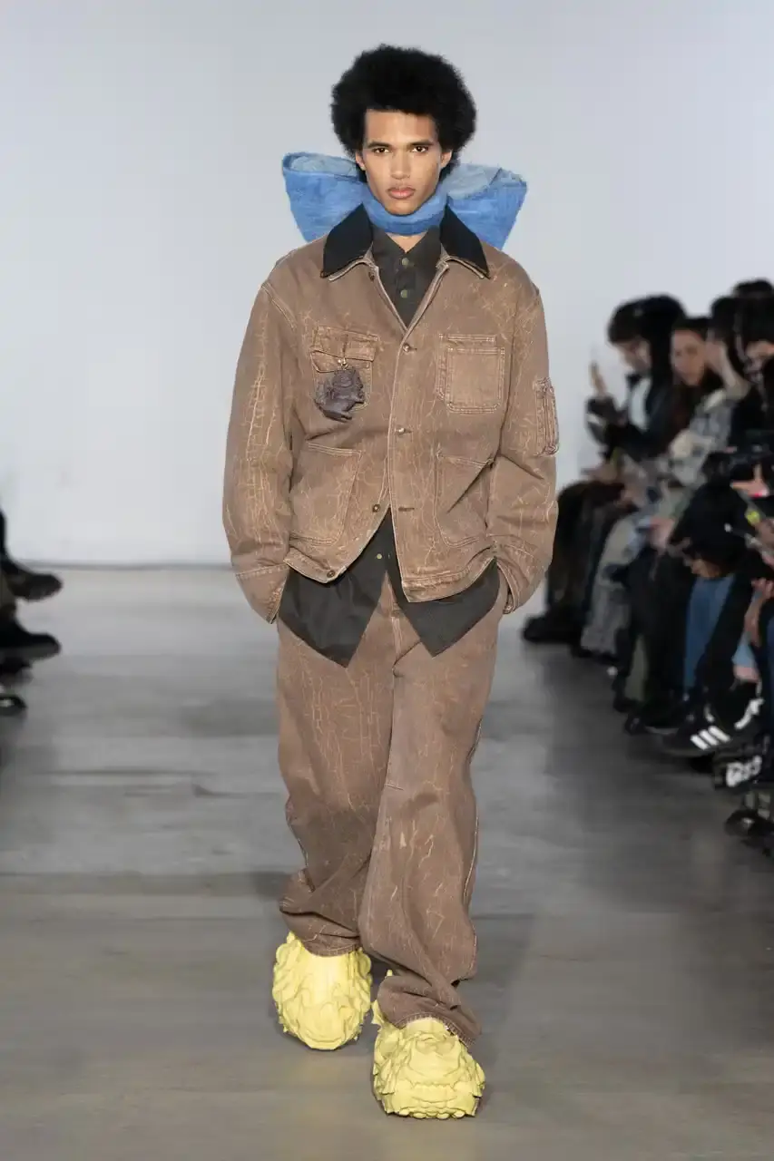 Feng Chen Wang Runway image of a model wearing Feng Chen Wang's Fall/Winter 2025 collection, showcasing oversized ceramic-textured UGG slippers and layered outerwear inspired by mythical Chinese creatures from the Shan Hai Jing text (image 2 of 11)