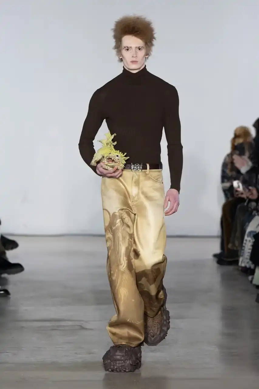 Feng Chen Wang Runway image of a model wearing Feng Chen Wang's Fall/Winter 2025 collection, showcasing oversized ceramic-textured UGG slippers and layered outerwear inspired by mythical Chinese creatures from the Shan Hai Jing text (image 10 of 11)