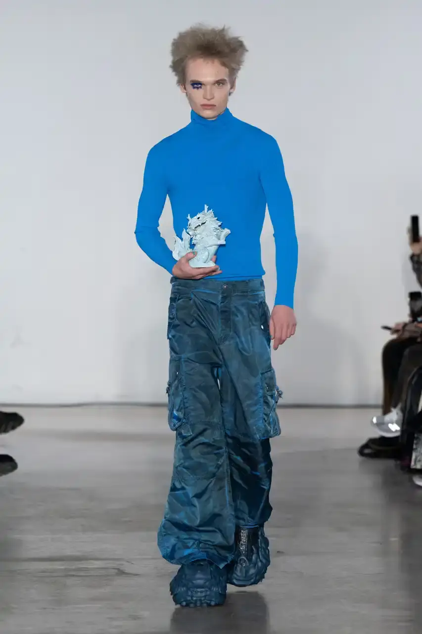 Feng Chen Wang Runway image of a model wearing Feng Chen Wang's Fall/Winter 2025 collection, showcasing oversized ceramic-textured UGG slippers and layered outerwear inspired by mythical Chinese creatures from the Shan Hai Jing text (image 4 of 11)