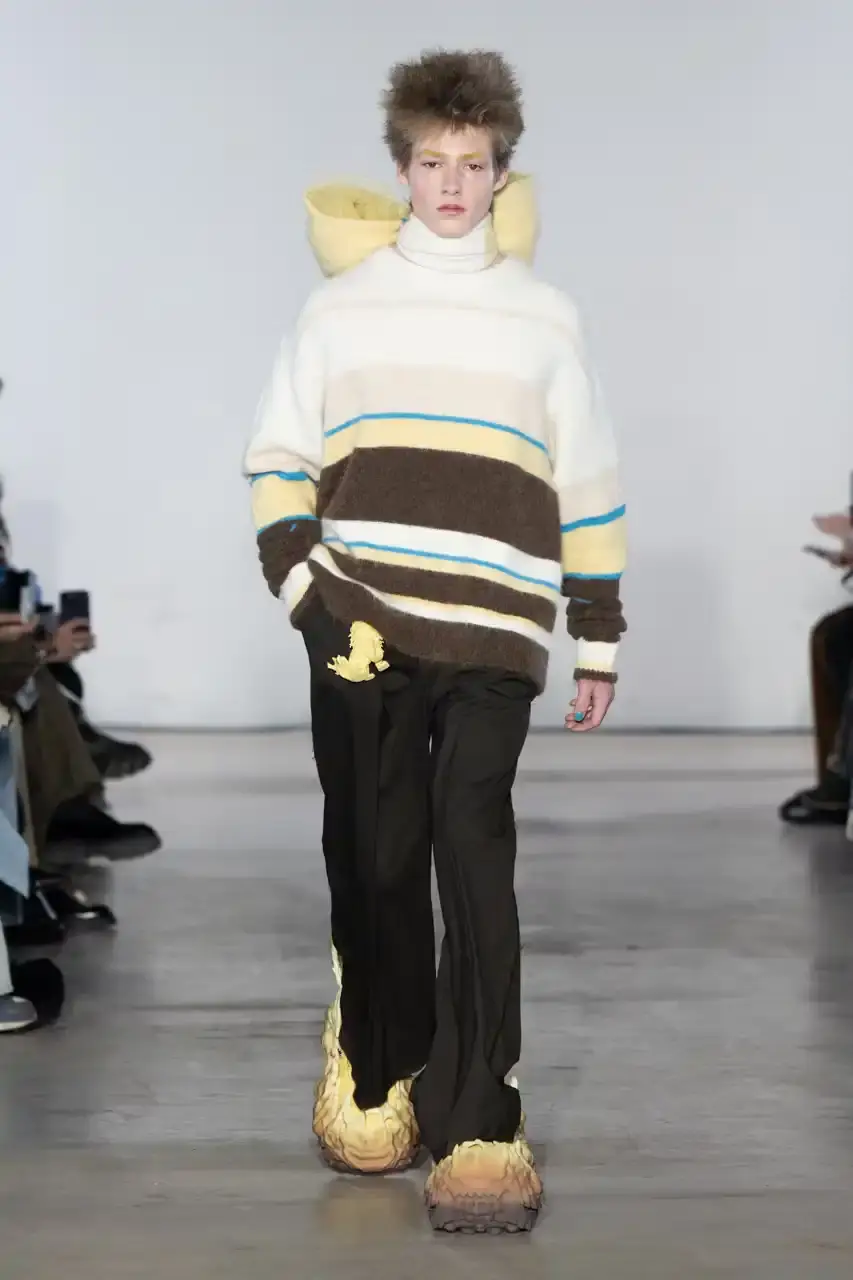 Feng Chen Wang Runway image of a model wearing Feng Chen Wang's Fall/Winter 2025 collection, showcasing oversized ceramic-textured UGG slippers and layered outerwear inspired by mythical Chinese creatures from the Shan Hai Jing text (image 8 of 11)