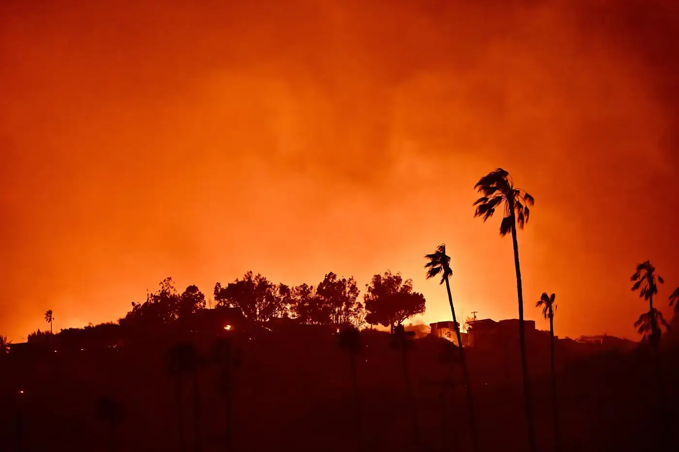Aerial view of wildfires burning in Los Angeles hills, with smoke billowing over the city skyline and residential areas in the foreground.