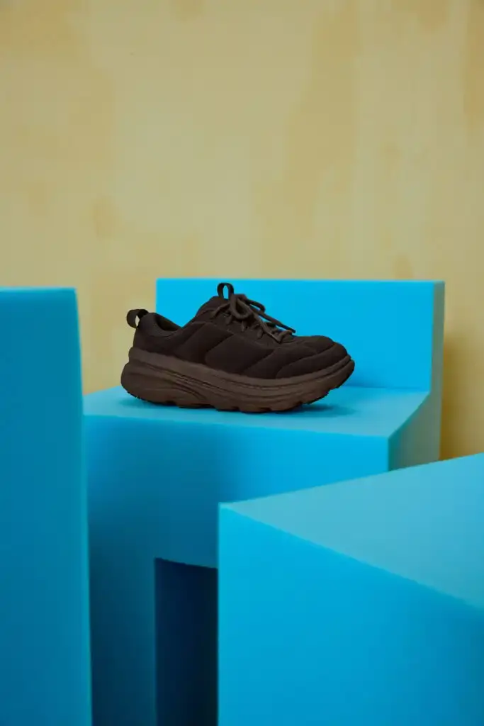 HOKA AND MARNI UNVEIL NEW SNEAKER PARTNERSHIP IN MILAN photo of the collaboration between athletic footwear brand Hoka and fashion label Marni on their new Bondi B3LS sneaker. It details the shoe's technical features including a cushioned sole, mesh upper with TPU reinforcements, and rubber outsole with traction lugs. The piece describes the sneaker's four color options (red, beige, blue, brown) and three interchangeable lace styles. The article also reports on the Milan launch event at Villa Clea featuring foam installations by Finemateria, performances by artist Marta Del Grandi and dancers Federica D'Aversa and Rafael Candela, plus a multisensory dining experience by Mimosa Milano. The sneakers are available for €395 on both brands' websites. (image 2 of 3)