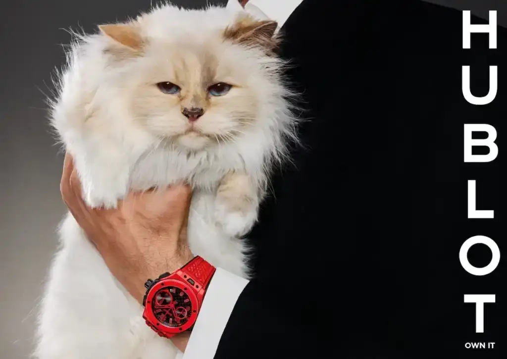 image of Hublot's 20th anniversary campaign for their Big Bang watch collection, featuring Karl Lagerfeld's cat Choupette as the unexpected star. Photographer Carlijn Jacobs captured both elegant editorial images and casual meme-inspired content centered on the Big Bang 20th Anniversary Red Magic timepiece. The campaign includes quotes from Hublot CEO Julien Tornare about breaking conventions in watchmaking, and uses the tagline "Own It" to reflect the brand's bold approach. The article explains how this campaign represents a departure from traditional luxury advertising by embracing playfulness and unpredictability on social media. (image 2 of 2)
