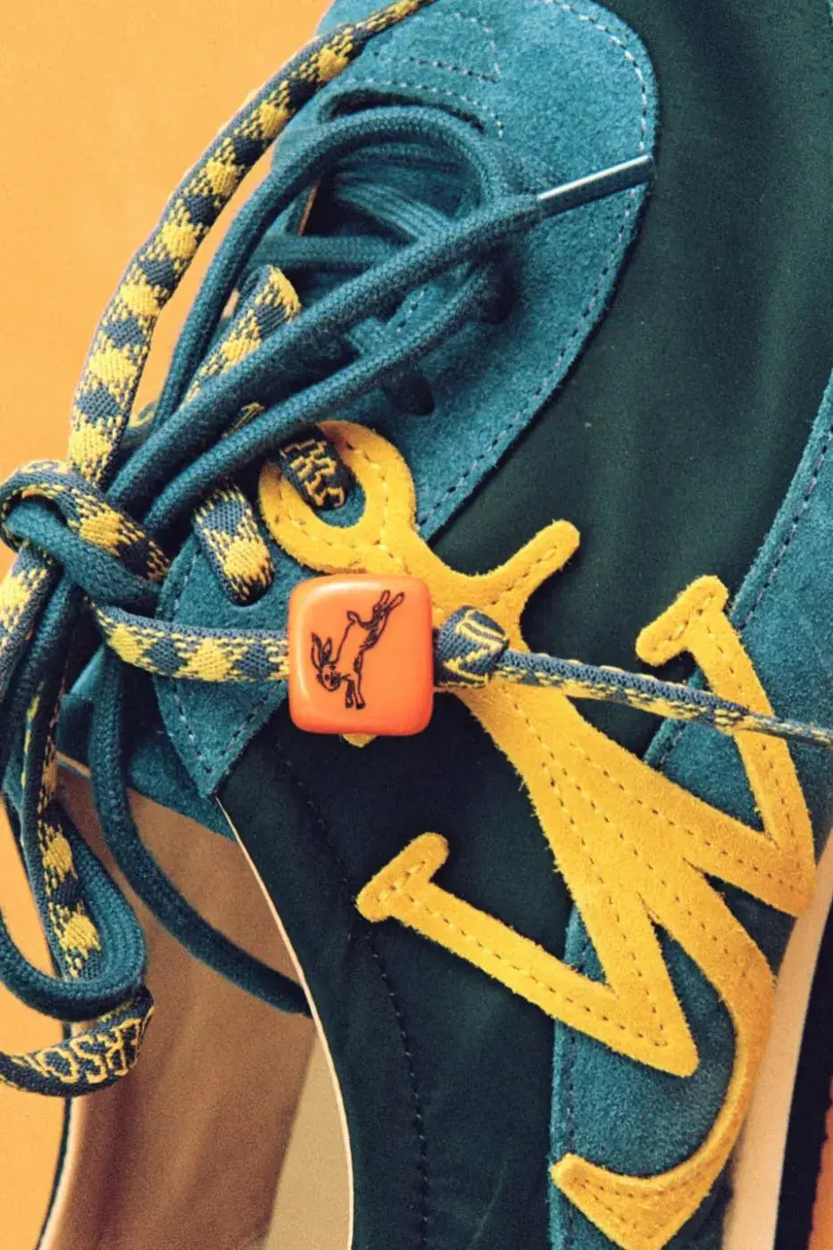 JW ANDERSON SHOWS THE “RUN RABBIT” TEAL SNEAKER image of the JW Anderson Run Rabbit sneaker with a running-shoe shape, a teal green suede-like upper, ochre yellow touches, patterned and rope-like laces and an orange dice-like charm with a running rabbit. (image 2 of 2)