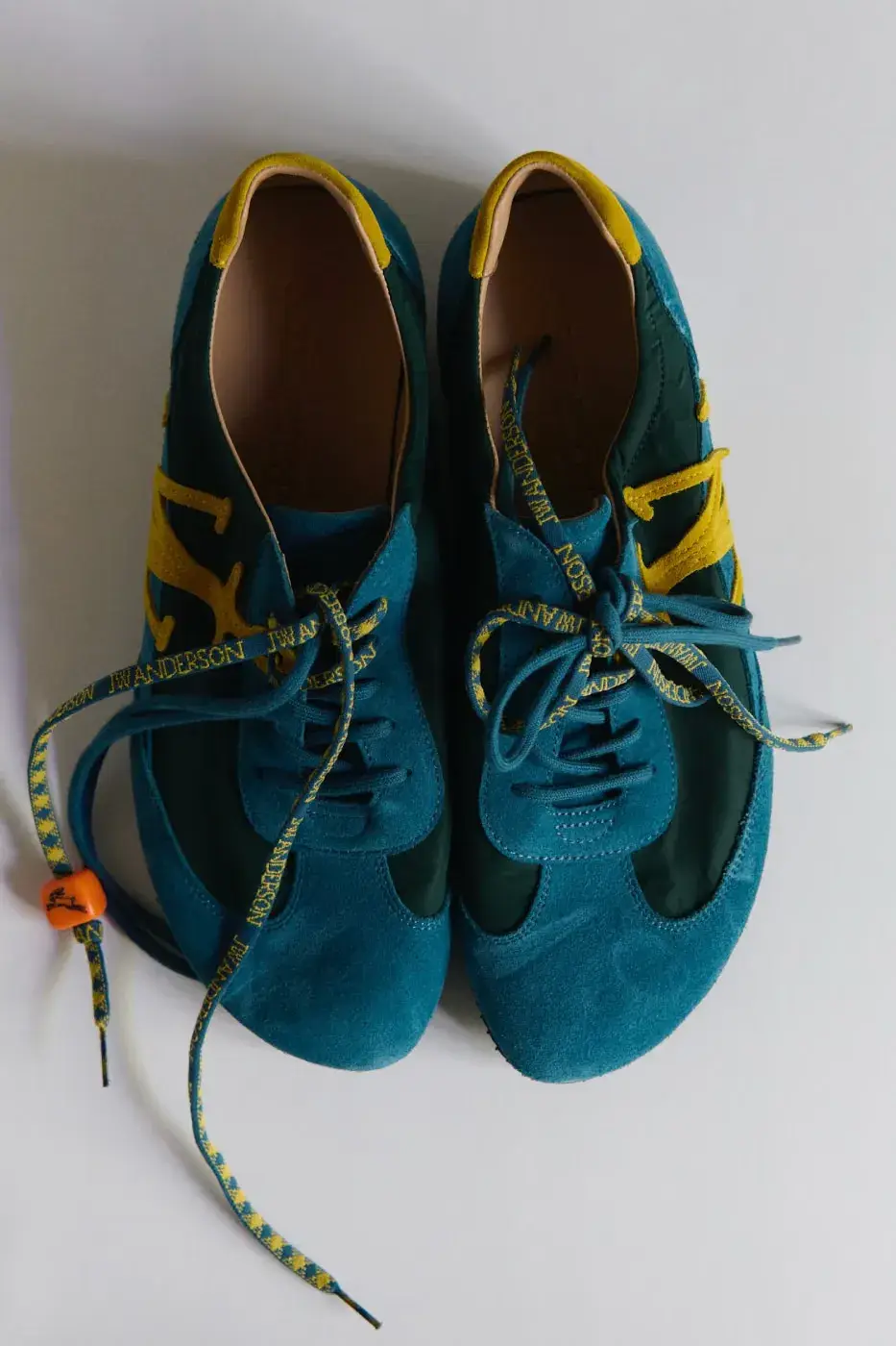 JW ANDERSON SHOWS THE “RUN RABBIT” TEAL SNEAKER image of the JW Anderson Run Rabbit sneaker with a running-shoe shape, a teal green suede-like upper, ochre yellow touches, patterned and rope-like laces and an orange dice-like charm with a running rabbit.