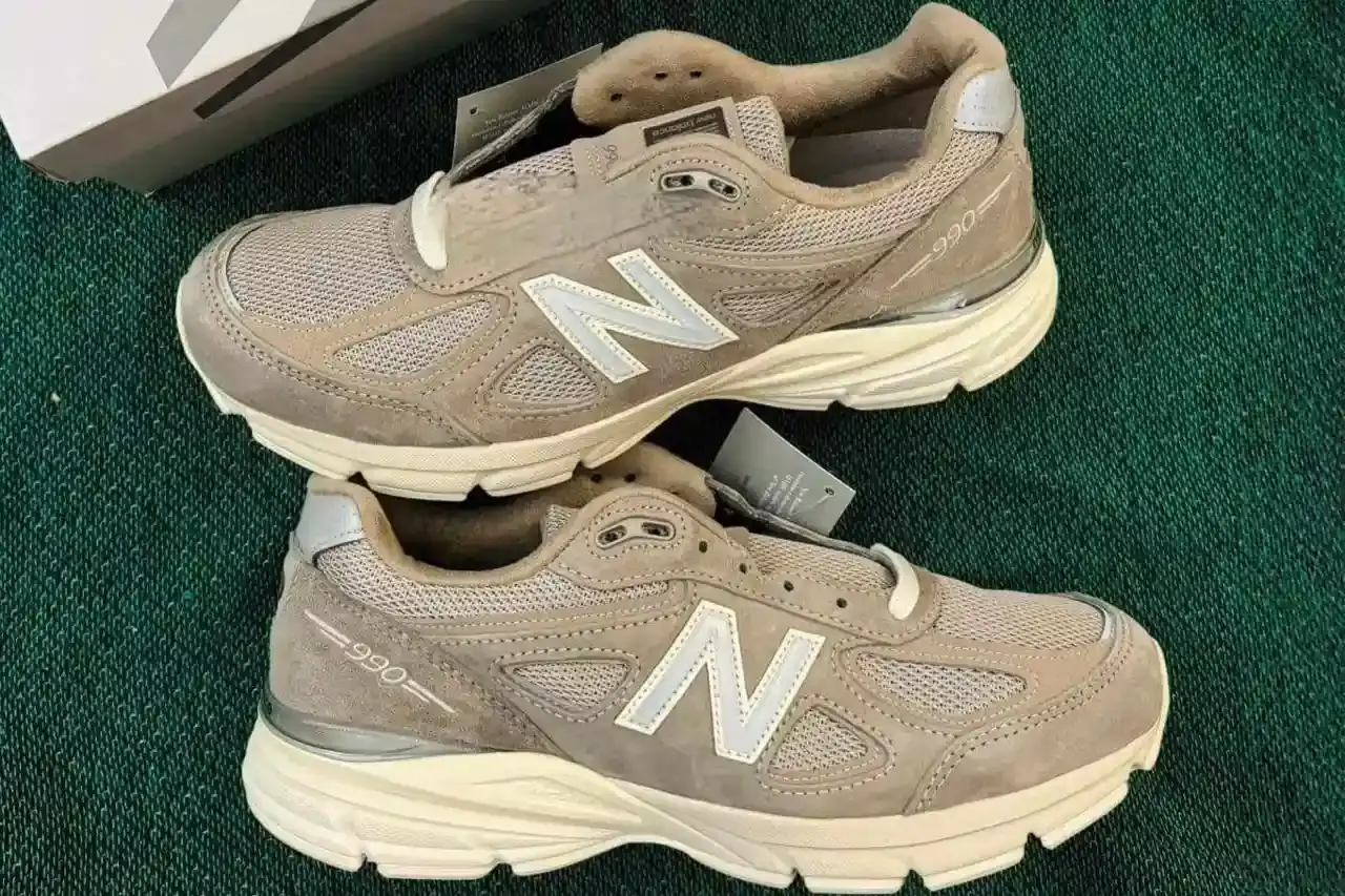 KITH x New Balance 990v4 sneaker in Grey and Cream colorway. Side view showing soft brown upper with grey 'N' logo, distinct KITH branding on heel, and off-white sole. A blend of classic design and contemporary streetwear aesthetics