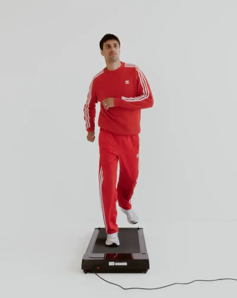 HOW SHAI DATAUKER REDEFINES CLOTHING, IDENTITY, AND DAILY LIFE Shai Datauker wearing his daily uniform-a bright red Adidas tracksuit and white t-shirt-sitting in a minimalist interior during an interview about fashion and identity. (image 4 of 5)