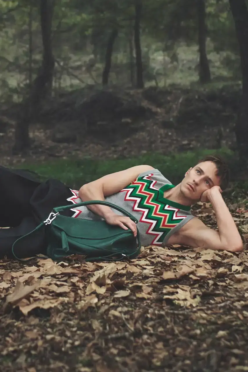 LOEWE Actor Drew Starkey reclines in tall mountain grass wearing a multicolored intarsia sweater vest and pleated trousers, with a green LOEWE puzzle bag beside him