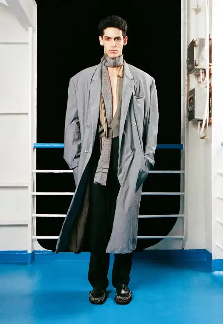 MAGLIANO SHOWS SS26 COLLECTION VIA FERRY FILM image about Italian designer Luca Magliano's Spring/Summer 2026 menswear collection, presented as a film aboard a ferry during Milan Fashion Week. The collection features camping and nomadic influences, with repurposed materials, loose silhouettes, and collaborative footwear including Beja soccer sneakers and 1900s-inspired boots. Shoot by Leonardo Scotti. (image 8 of 16)