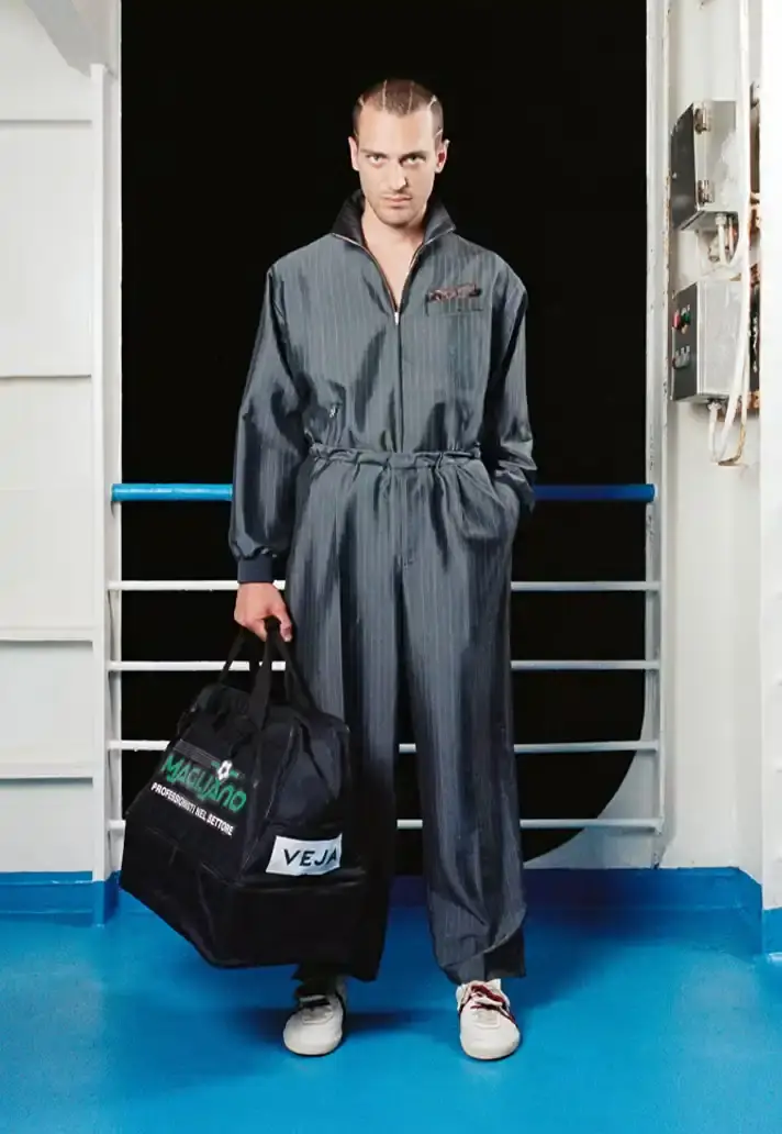 MAGLIANO SHOWS SS26 COLLECTION VIA FERRY FILM image about Italian designer Luca Magliano's Spring/Summer 2026 menswear collection, presented as a film aboard a ferry during Milan Fashion Week. The collection features camping and nomadic influences, with repurposed materials, loose silhouettes, and collaborative footwear including Beja soccer sneakers and 1900s-inspired boots. Shoot by Leonardo Scotti. (image 14 of 16)
